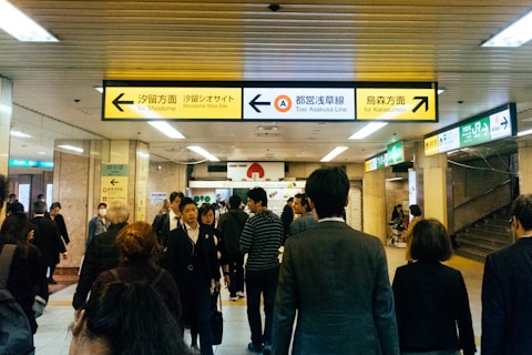 A crowded indoor scene depicting a subway station with multiple people walking in various directions. The ceiling has fluorescent lighting and there are yellow directional signs pointing to different subway lines and destinations such as 'Shiodome' and 'Karasumori'. The walls of the station are lined with various signs and notices. The people are dressed in a mix of casual and business attire, some wearing masks.