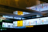 A series of colorful wayfinding signs, featuring directions and line information for various train services. The signs include both English and Japanese text, with symbols indicating facilities such as restrooms and accessible features. There's a notable reflection of the signage on the ceiling, adding depth to the image.