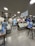 A modern eyewear store with several display tables featuring a variety of eyeglasses. The store has a sleek design with bright ceiling lights, digital screens, and branded displays. Customers are browsing and trying on glasses. A sign offering a free eye test hangs from the ceiling.