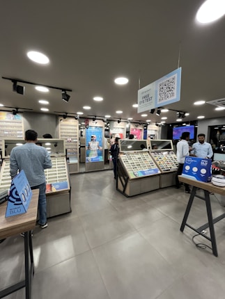 A modern eyewear store with several display tables featuring a variety of eyeglasses. The store has a sleek design with bright ceiling lights, digital screens, and branded displays. Customers are browsing and trying on glasses. A sign offering a free eye test hangs from the ceiling.