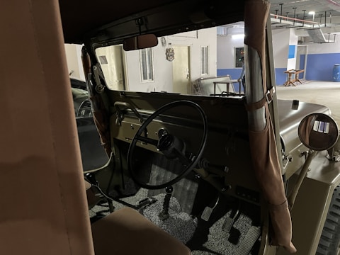 The interior view of an old military-style vehicle or jeep is visible, with a focus on the steering wheel, dashboard, and pedals. The environment seems to be a dimly lit parking area or garage with concrete walls and visible ceiling structures. There are hints of natural and artificial lighting.