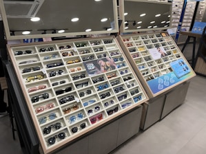 A display case filled with a variety of sunglasses, each positioned in neat rows and columns within their respective compartments. The display is well-lit, with overhead round lights reflecting off shiny surfaces. The case includes promotional images and informational signs about the sunglasses. Mirrors above the display add depth and space to the scene.