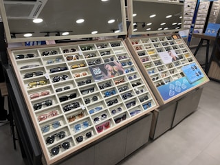 A display case filled with a variety of sunglasses, each positioned in neat rows and columns within their respective compartments. The display is well-lit, with overhead round lights reflecting off shiny surfaces. The case includes promotional images and informational signs about the sunglasses. Mirrors above the display add depth and space to the scene.