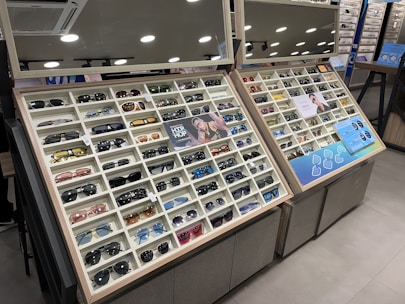 A display case filled with a variety of sunglasses, each positioned in neat rows and columns within their respective compartments. The display is well-lit, with overhead round lights reflecting off shiny surfaces. The case includes promotional images and informational signs about the sunglasses. Mirrors above the display add depth and space to the scene.