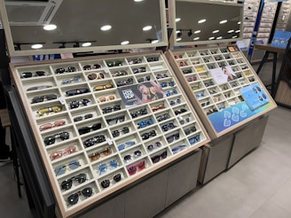 A display case filled with a variety of sunglasses, each positioned in neat rows and columns within their respective compartments. The display is well-lit, with overhead round lights reflecting off shiny surfaces. The case includes promotional images and informational signs about the sunglasses. Mirrors above the display add depth and space to the scene.