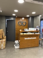 A retail store interior featuring a wooden customer service desk with the logo of an eyewear company 'lenskart.com' prominently displayed. Several small stacks of product boxes and informational pamphlets are placed on the desk. Behind the desk, a wall with a wood panel design displays a large eyeglasses logo. The lighting is bright with recessed ceiling lights, and there are security cameras visible. On the floor, a few cardboard boxes are stacked to the side.