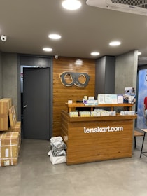 A retail store interior featuring a wooden customer service desk with the logo of an eyewear company 'lenskart.com' prominently displayed. Several small stacks of product boxes and informational pamphlets are placed on the desk. Behind the desk, a wall with a wood panel design displays a large eyeglasses logo. The lighting is bright with recessed ceiling lights, and there are security cameras visible. On the floor, a few cardboard boxes are stacked to the side.