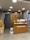 A retail store interior featuring a wooden customer service desk with the logo of an eyewear company 'lenskart.com' prominently displayed. Several small stacks of product boxes and informational pamphlets are placed on the desk. Behind the desk, a wall with a wood panel design displays a large eyeglasses logo. The lighting is bright with recessed ceiling lights, and there are security cameras visible. On the floor, a few cardboard boxes are stacked to the side.