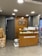 A retail store interior featuring a wooden customer service desk with the logo of an eyewear company 'lenskart.com' prominently displayed. Several small stacks of product boxes and informational pamphlets are placed on the desk. Behind the desk, a wall with a wood panel design displays a large eyeglasses logo. The lighting is bright with recessed ceiling lights, and there are security cameras visible. On the floor, a few cardboard boxes are stacked to the side.
