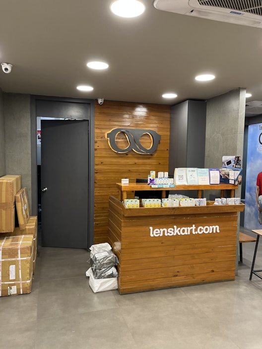 A retail store interior featuring a wooden customer service desk with the logo of an eyewear company 'lenskart.com' prominently displayed. Several small stacks of product boxes and informational pamphlets are placed on the desk. Behind the desk, a wall with a wood panel design displays a large eyeglasses logo. The lighting is bright with recessed ceiling lights, and there are security cameras visible. On the floor, a few cardboard boxes are stacked to the side.
