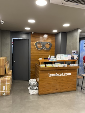 A retail store interior featuring a wooden customer service desk with the logo of an eyewear company 'lenskart.com' prominently displayed. Several small stacks of product boxes and informational pamphlets are placed on the desk. Behind the desk, a wall with a wood panel design displays a large eyeglasses logo. The lighting is bright with recessed ceiling lights, and there are security cameras visible. On the floor, a few cardboard boxes are stacked to the side.
