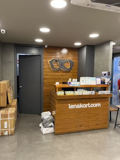 A retail store interior featuring a wooden customer service desk with the logo of an eyewear company 'lenskart.com' prominently displayed. Several small stacks of product boxes and informational pamphlets are placed on the desk. Behind the desk, a wall with a wood panel design displays a large eyeglasses logo. The lighting is bright with recessed ceiling lights, and there are security cameras visible. On the floor, a few cardboard boxes are stacked to the side.