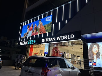 The image depicts the exterior of a 'Titan World' store at night, featuring bright signage and illuminated displays. Several advertisements are visible, including a large digital banner showcasing models on a blue background and a display window with fashion imagery. A car and a motorbike are parked in front of the store.
