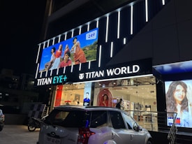 The image depicts the exterior of a 'Titan World' store at night, featuring bright signage and illuminated displays. Several advertisements are visible, including a large digital banner showcasing models on a blue background and a display window with fashion imagery. A car and a motorbike are parked in front of the store.