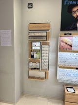 A display wall featuring multiple shelves with various eyeglasses and related accessories. The shelves are labeled 'ACCESSORIES' and include categories like 'COMPUTER GLASSES', 'READY READERS', and 'CLIP-ON FRAMES'. There are promotional materials and images of glasses on the wall, as well as a signboard thanking customers for shopping. A sign indicates the store is under CCTV surveillance.