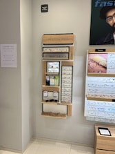 A display wall featuring multiple shelves with various eyeglasses and related accessories. The shelves are labeled 'ACCESSORIES' and include categories like 'COMPUTER GLASSES', 'READY READERS', and 'CLIP-ON FRAMES'. There are promotional materials and images of glasses on the wall, as well as a signboard thanking customers for shopping. A sign indicates the store is under CCTV surveillance.