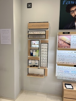A display wall featuring multiple shelves with various eyeglasses and related accessories. The shelves are labeled 'ACCESSORIES' and include categories like 'COMPUTER GLASSES', 'READY READERS', and 'CLIP-ON FRAMES'. There are promotional materials and images of glasses on the wall, as well as a signboard thanking customers for shopping. A sign indicates the store is under CCTV surveillance.