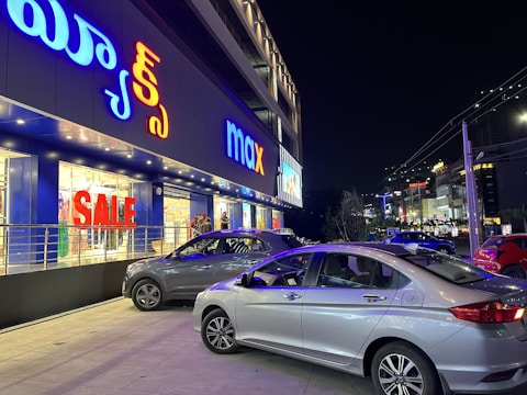A busy street scene at night featuring a brightly lit storefront with signage in multiple languages. Large 'SALE' signs are visible through the windows. Several parked cars occupy the foreground, and additional storefronts with illuminated signs are visible in the background.