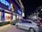 A busy street scene at night featuring a brightly lit storefront with signage in multiple languages. Large 'SALE' signs are visible through the windows. Several parked cars occupy the foreground, and additional storefronts with illuminated signs are visible in the background.