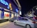 A busy street scene at night featuring a brightly lit storefront with signage in multiple languages. Large 'SALE' signs are visible through the windows. Several parked cars occupy the foreground, and additional storefronts with illuminated signs are visible in the background.
