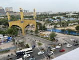 A busy intersection features numerous vehicles including cars, buses, and motorcycles. A large golden arch with the word 'HITEX' stands prominently on one side of the road. Trees and greenery are visible in the background, along with high-rise buildings and blue fencing. People can be seen moving around the area on foot or using vehicles. Several advertisements and signs are present, adding to the urban environment.
