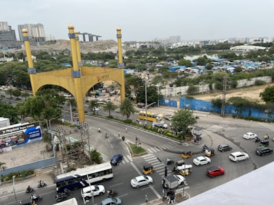 A busy intersection features numerous vehicles including cars, buses, and motorcycles. A large golden arch with the word 'HITEX' stands prominently on one side of the road. Trees and greenery are visible in the background, along with high-rise buildings and blue fencing. People can be seen moving around the area on foot or using vehicles. Several advertisements and signs are present, adding to the urban environment.