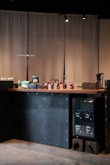 A minimalist coffee shop counter with a wooden countertop displaying various items including bags of coffee, cups, and jars. To the right, a small glass-front refrigerator contains labeled items. The backdrop consists of beige curtains, and the lighting is soft and focused.