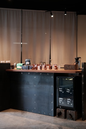 A minimalist coffee shop counter with a wooden countertop displaying various items including bags of coffee, cups, and jars. To the right, a small glass-front refrigerator contains labeled items. The backdrop consists of beige curtains, and the lighting is soft and focused.