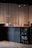 A minimalist coffee shop counter with a wooden countertop displaying various items including bags of coffee, cups, and jars. To the right, a small glass-front refrigerator contains labeled items. The backdrop consists of beige curtains, and the lighting is soft and focused.