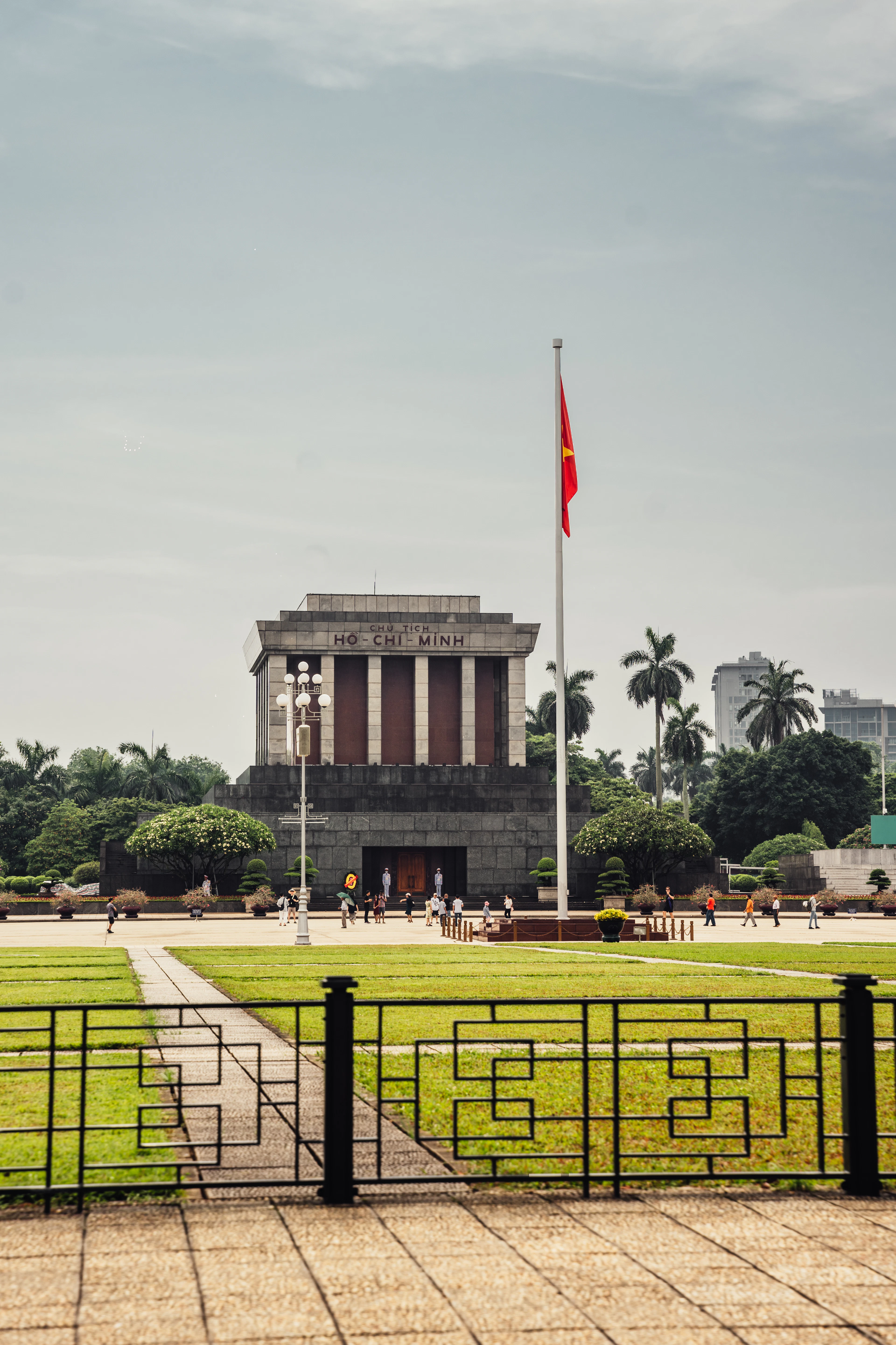 The Ho Chi Minh Mausoleum stands under blue sky with the Vietnamese flag flying above it.