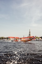 A colorful long-tail boat moves swiftly across a river with splashes of water in the foreground. In the background, there is a scenic view of a traditional Asian temple with a tall, ornate spire rising into the clear sky. The riverbank is adorned with greenery and red-roofed buildings.
