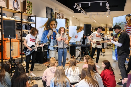 A small band is performing in a retail clothing store. The musicians are playing various instruments surrounded by racks of denim jeans and promotional posters. A group of people sit on the floor, watching the performance attentively. There's a lively, casual atmosphere with a mix of music and shopping.