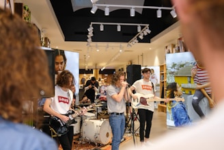 A band is performing in a store, featuring a guitarist, a singer, and a bass player, with two large screens displaying images behind them. People are casually watching the performance in a modern retail environment, and the store appears to have a high ceiling with track lighting.