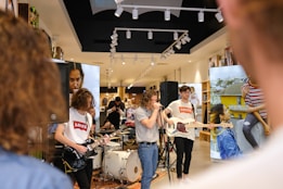 A band is performing in a store, featuring a guitarist, a singer, and a bass player, with two large screens displaying images behind them. People are casually watching the performance in a modern retail environment, and the store appears to have a high ceiling with track lighting.