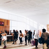 A group of people stand in line inside a bright and spacious building with large windows. The sunlight creates shadows on the floor, and some individuals carry luggage and wear backpacks. There is a counter with staff assisting the individuals. The interior features wood paneling and minimalistic decor.