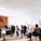 A group of people stand in line inside a bright and spacious building with large windows. The sunlight creates shadows on the floor, and some individuals carry luggage and wear backpacks. There is a counter with staff assisting the individuals. The interior features wood paneling and minimalistic decor.