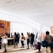 A group of people stand in line inside a bright and spacious building with large windows. The sunlight creates shadows on the floor, and some individuals carry luggage and wear backpacks. There is a counter with staff assisting the individuals. The interior features wood paneling and minimalistic decor.