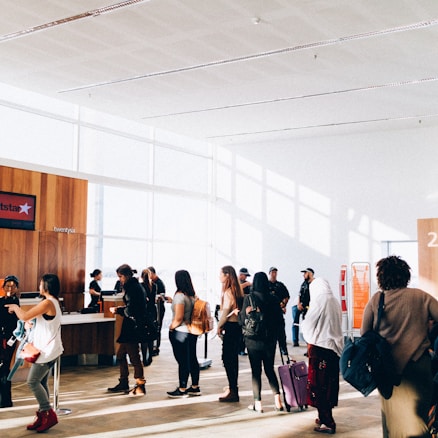 A group of people stand in line inside a bright and spacious building with large windows. The sunlight creates shadows on the floor, and some individuals carry luggage and wear backpacks. There is a counter with staff assisting the individuals. The interior features wood paneling and minimalistic decor.