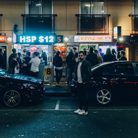 A bustling street scene at night in front of a kebab shop with a large crowd of people waiting and mingling outside. Bright neon signs and window lights illuminate the area. Several parked cars are visible, and a person stands prominently in the foreground.