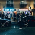 A bustling street scene at night in front of a kebab shop with a large crowd of people waiting and mingling outside. Bright neon signs and window lights illuminate the area. Several parked cars are visible, and a person stands prominently in the foreground.