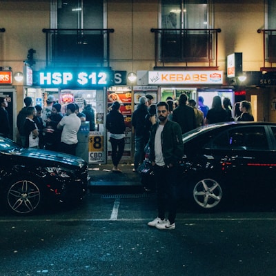 A bustling street scene at night in front of a kebab shop with a large crowd of people waiting and mingling outside. Bright neon signs and window lights illuminate the area. Several parked cars are visible, and a person stands prominently in the foreground.