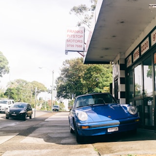 A vintage blue Porsche is parked next to a small auto repair shop with a sign that reads 'Franks Pitstop Motors.' The setting is a quiet suburban street with a few cars passing by. The building has a black and white striped exterior near the entrance, and trees are visible in the background.