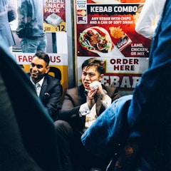 A group of men dressed in formal suits are sitting near a fast-food kiosk displaying colorful signs advertising kebabs. One man holds a wrapped kebab, partially eaten, and appears to be speaking animatedly. The setting is lively, with bright signage and the presence of other people in casual attire nearby.