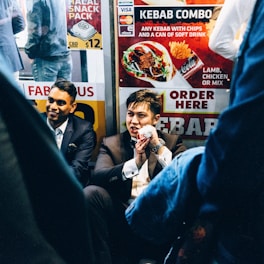 A group of men dressed in formal suits are sitting near a fast-food kiosk displaying colorful signs advertising kebabs. One man holds a wrapped kebab, partially eaten, and appears to be speaking animatedly. The setting is lively, with bright signage and the presence of other people in casual attire nearby.