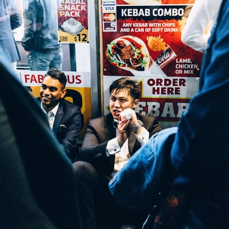 A group of men dressed in formal suits are sitting near a fast-food kiosk displaying colorful signs advertising kebabs. One man holds a wrapped kebab, partially eaten, and appears to be speaking animatedly. The setting is lively, with bright signage and the presence of other people in casual attire nearby.