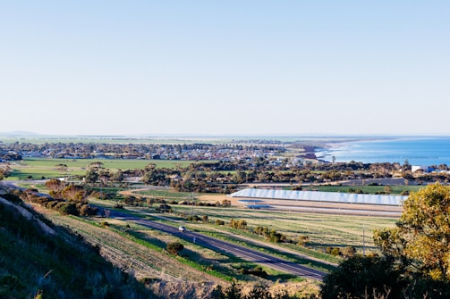A sprawling landscape features a coastline with turquoise waters meeting the sky at the horizon. In the foreground, open fields and roads are visible, with scattered buildings and a large greenhouse structure. The distant view includes a small town or settlement with clusters of trees providing some greenery.