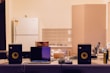 A kitchen counter is cluttered with various electronic devices and items. Two black speakers are positioned on either side of an open laptop. A box of tea sits next to the laptop, along with tangled cables. In the background, a white refrigerator and cabinets are visible against a tiled wall.