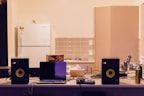 A kitchen counter is cluttered with various electronic devices and items. Two black speakers are positioned on either side of an open laptop. A box of tea sits next to the laptop, along with tangled cables. In the background, a white refrigerator and cabinets are visible against a tiled wall.