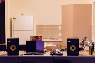 A kitchen counter is cluttered with various electronic devices and items. Two black speakers are positioned on either side of an open laptop. A box of tea sits next to the laptop, along with tangled cables. In the background, a white refrigerator and cabinets are visible against a tiled wall.