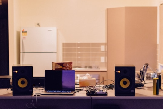 A kitchen counter is cluttered with various electronic devices and items. Two black speakers are positioned on either side of an open laptop. A box of tea sits next to the laptop, along with tangled cables. In the background, a white refrigerator and cabinets are visible against a tiled wall.
